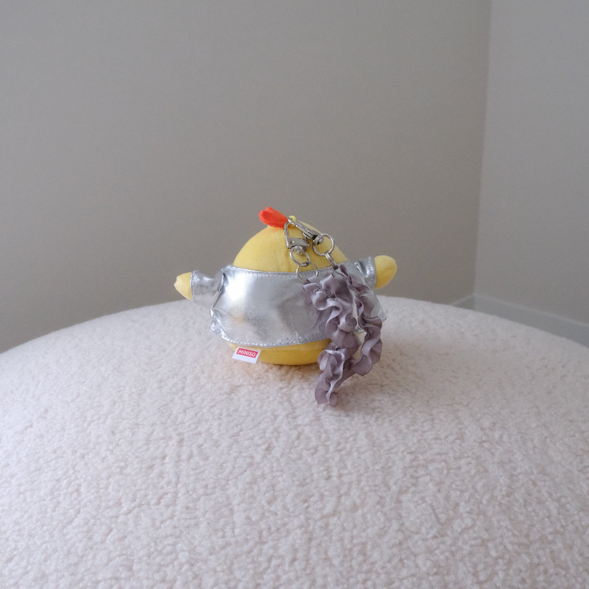 A small yellow plush toy wearing a silver metallic shirt and a ruffled lavender-gray ruffled wristlet strap, resting on a textured cream-colored surface against a plain light gray wall.