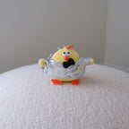 A small yellow chick figurine wearing a shiny silver suit, standing on a textured white surface against a plain light gray wall.