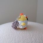 A small plush chicken keychain with a yellow body, orange crown and feet, black beak, and a silver foil top, standing on a textured white surface against a plain light gray wall.