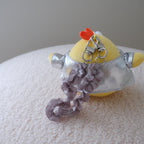 A small plush toy or accessory with a yellow rounded body, silver metallic-top , a gray ruffled wristlet strap, and a small orange chicken crown on top, photographed resting on a white textured surface against a light gray background.