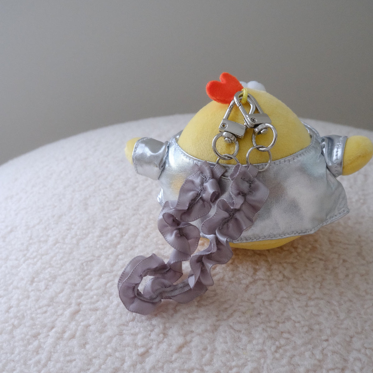 A small plush toy or accessory with a yellow rounded body, silver metallic-top , a gray ruffled wristlet strap, and a small orange chicken crown on top, photographed resting on a white textured surface against a light gray background.